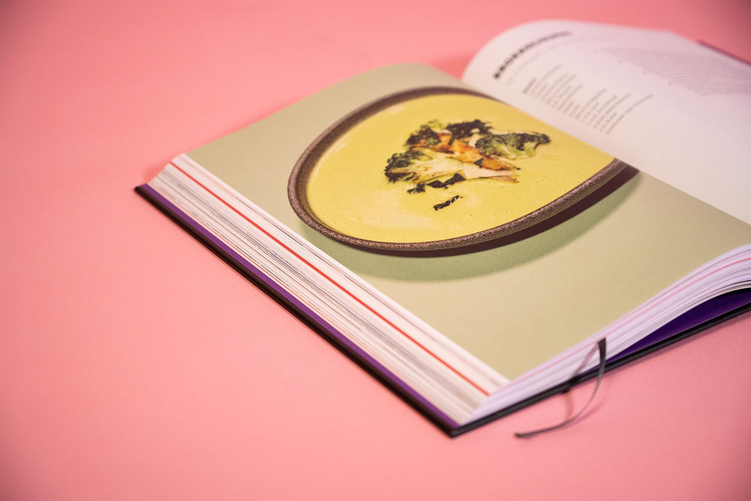 Buch aufgeschlagen. Seite mit Gericht. Auf rosa Untergrund. Gericht auf Teller.