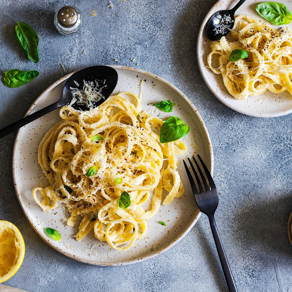 Zwei Teller Pasta mit Käse und Basilikum. Schwarze Löffel und Gabel. Zitrone und Gewürzstreuer.