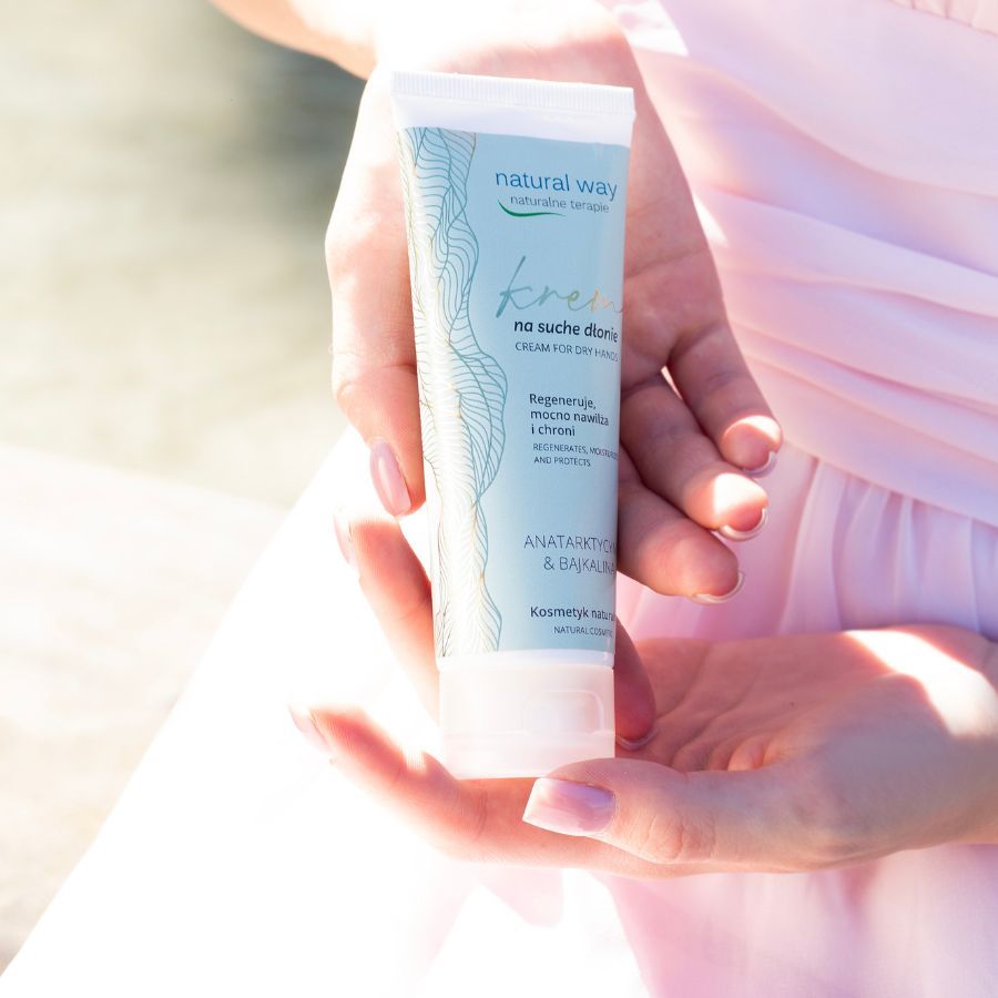 Creme-Tube, gehalten von Händen. Aufschrift: Natural Way, Creme für trockene Hände. Rosa Hintergrund.