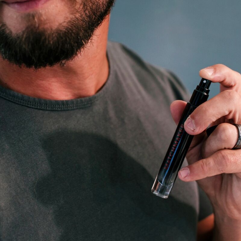 Nahaufnahme eines Mannes, der Parfüm aufsprüht. Schwarzer Flakon mit roter Schrift. Der Mann trägt ein graues T-Shirt.