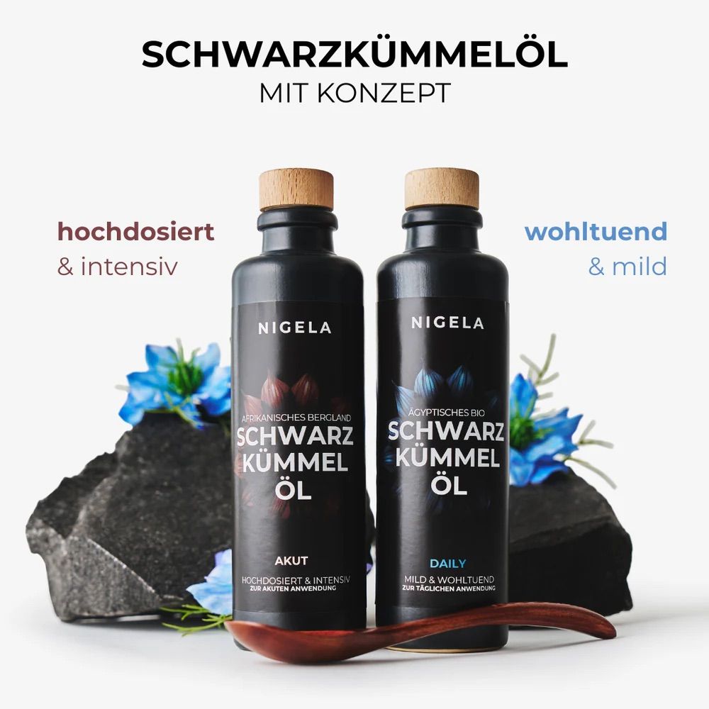 Zwei Flaschen Nigella Schwarzkümmelöl (AKUT & DAILY) mit Holzverschluss, auf Steinen mit blauen Blüten und Holzlöffel.