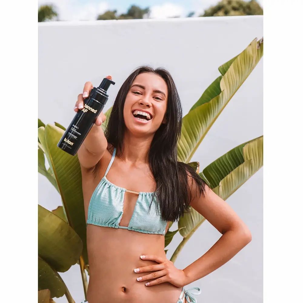 Frau hält eine schwarze Flasche 'tanned AF' und 'b.tan' in die Kamera. Bikini. Hintergrund: Pflanzen.