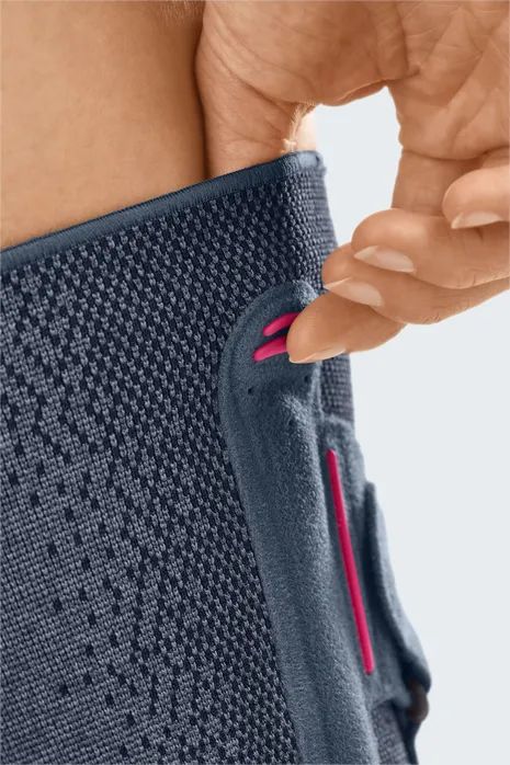 Nahaufnahme der Knieorthese. Dunkelgraues, gestricktes Material. Pinkfarbene Details. Hand berührt das Produkt.