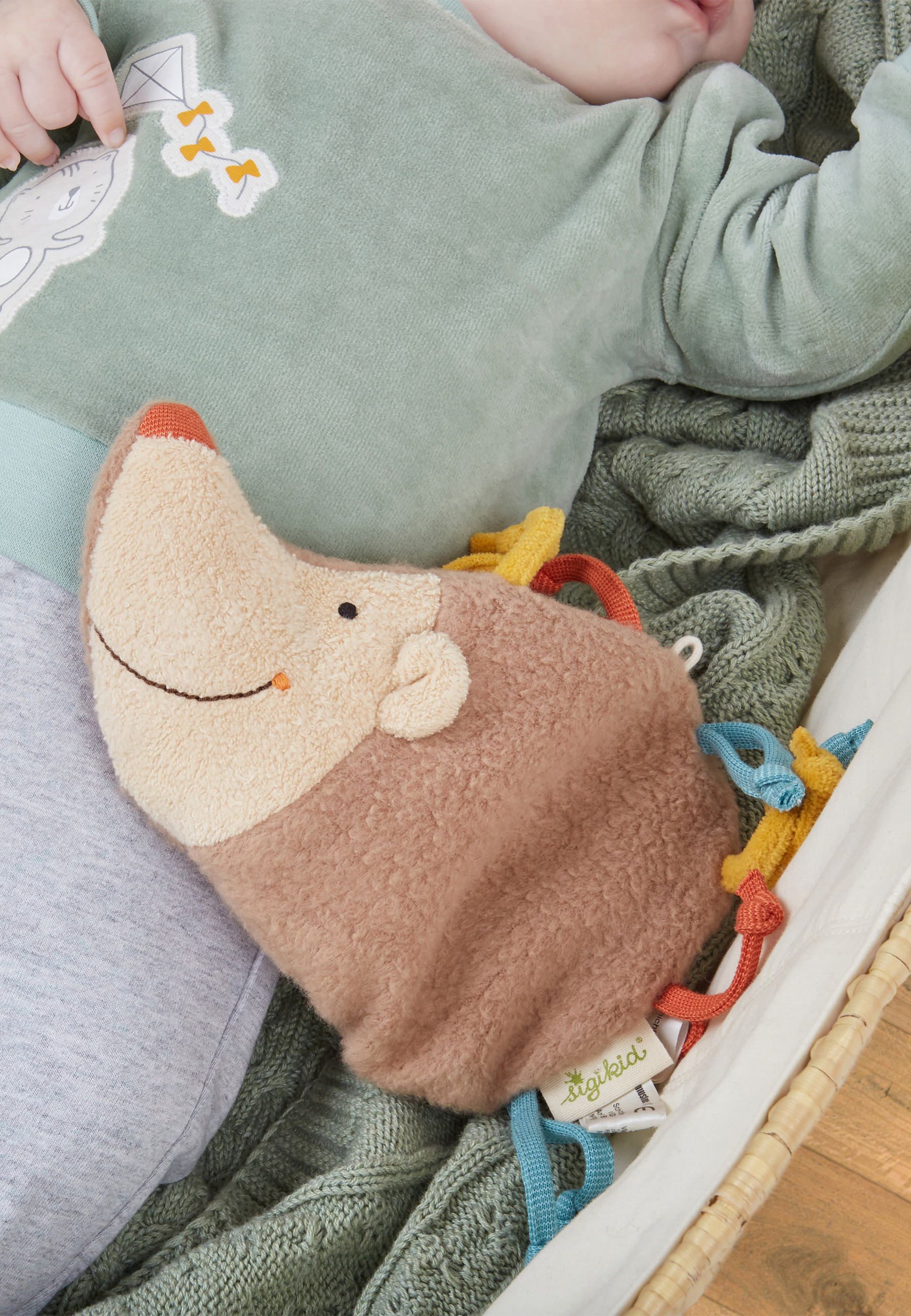 Baby liegt neben Stofftier-Igel. Igel mit bunten Bändern. Beige-braune Farbe, roter Mund. Marke SIGIKID.