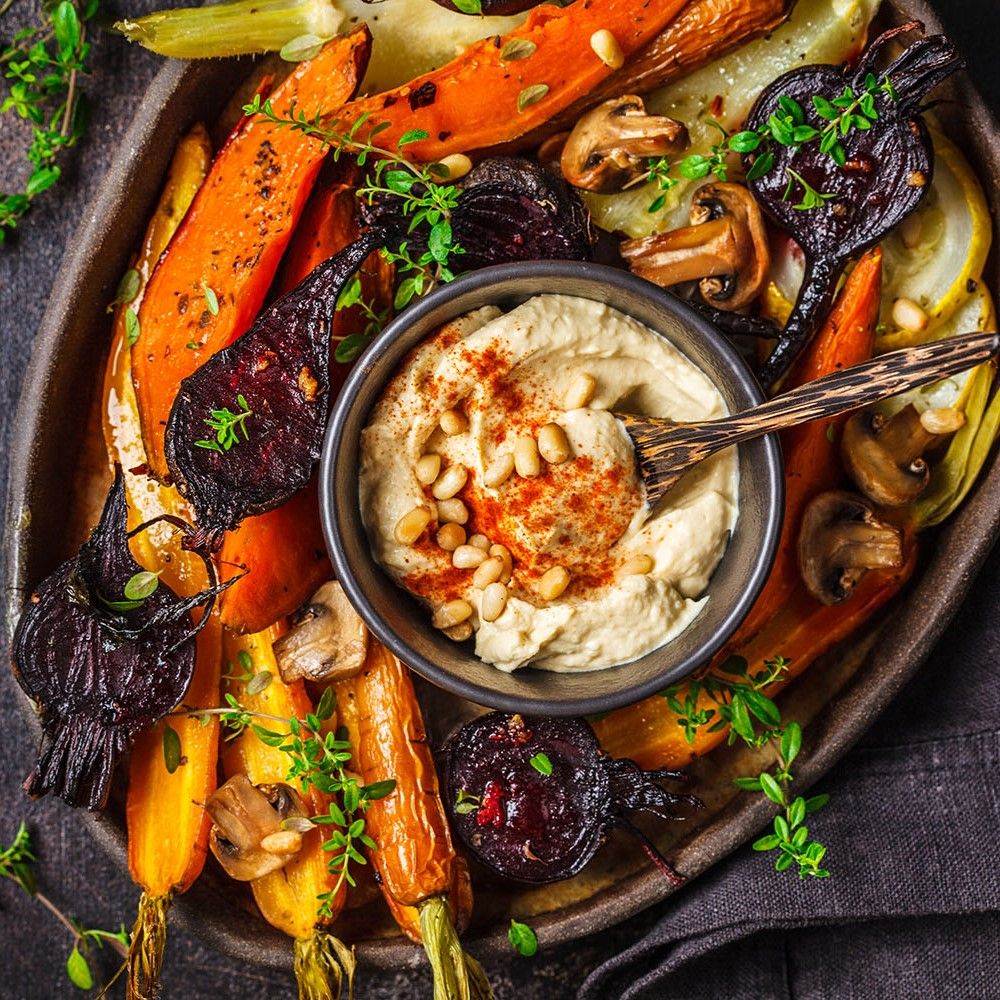 Hummus in Schüssel, umgeben von geröstetem Gemüse. Karotten, Pilze, Rote Bete. Auf dunklem Untergrund.
