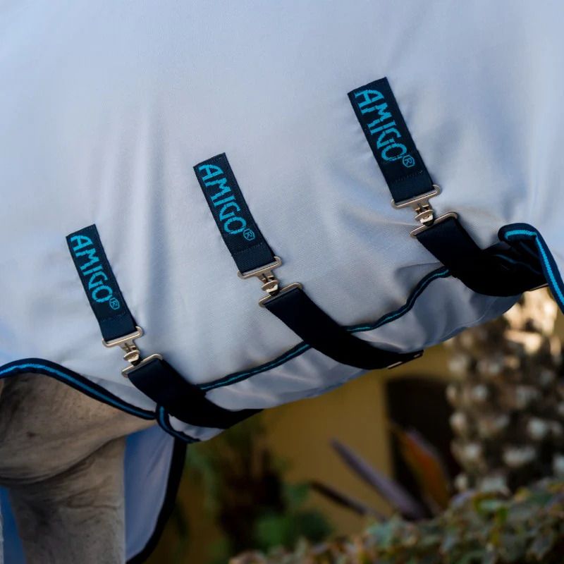 Nahaufnahme von drei schwarzen Gurten mit Metallverschlüssen an einer hellblauen Pferdedecke. Auf den Gurten steht 'AMIGO'.