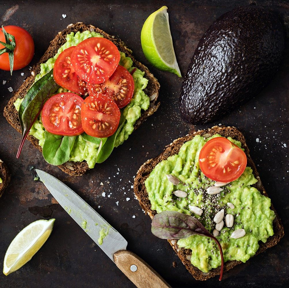 Zwei Brotscheiben mit Avocado-Creme, Tomaten und Sonnenblumenkernen. Limetten und Tomaten daneben. Messer.