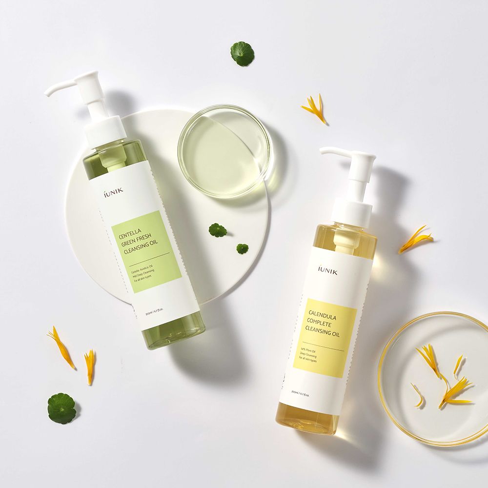 Zwei Flaschen iUNIK Cleansing Oil. Eine grün, eine gelb. Auf weißem Teller. Blütenblätter und grüne Blätter daneben.