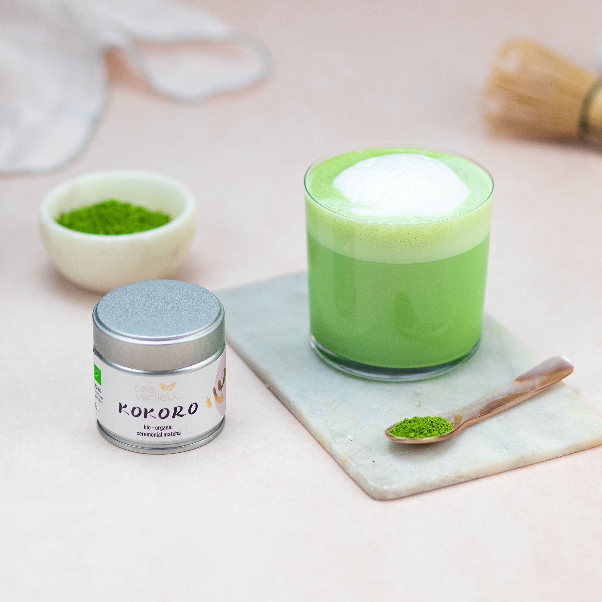 Glas Matcha-Getränk mit Schaum. Dose "teeverliebt Kokoro" und Schale mit Matcha. Löffel mit Matcha. Bambusbesen.