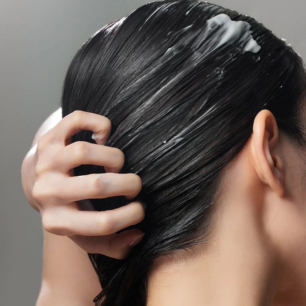 Person mit dunklem Haar. Weiße Creme auf dem Haar. Hand massiert das Haar. Grauer Hintergrund.