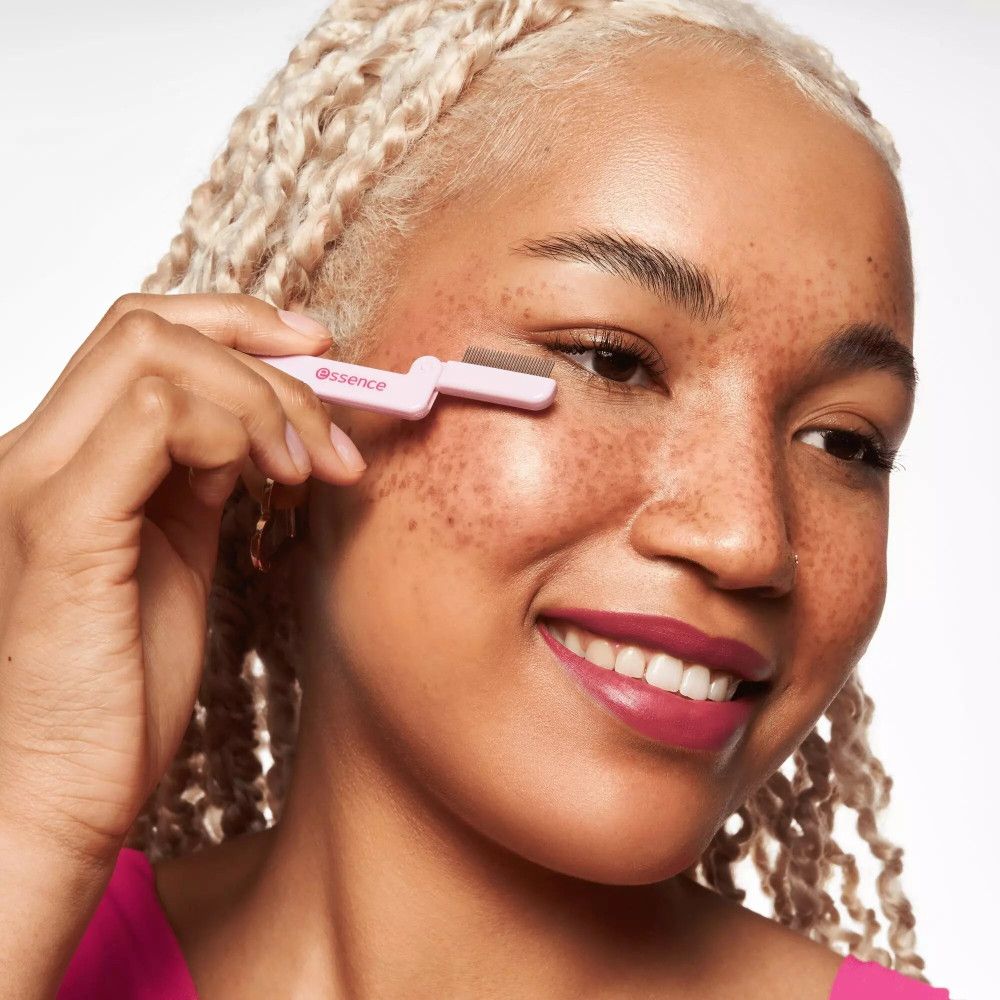 Person hält einen rosa Wimpernkamm. Marke: Essence. Die Person lächelt. Der Kamm wird am Auge verwendet.