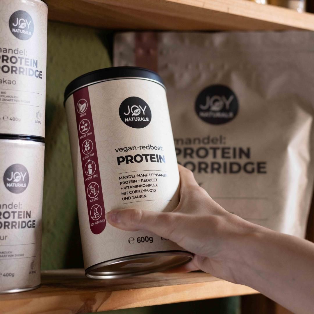Person hält Dose JOY NATURALS vegan-redbeet:PROTEIN. Aufschrift: Protein, 600g. Weitere Dosen und Beutel im Hintergrund.
