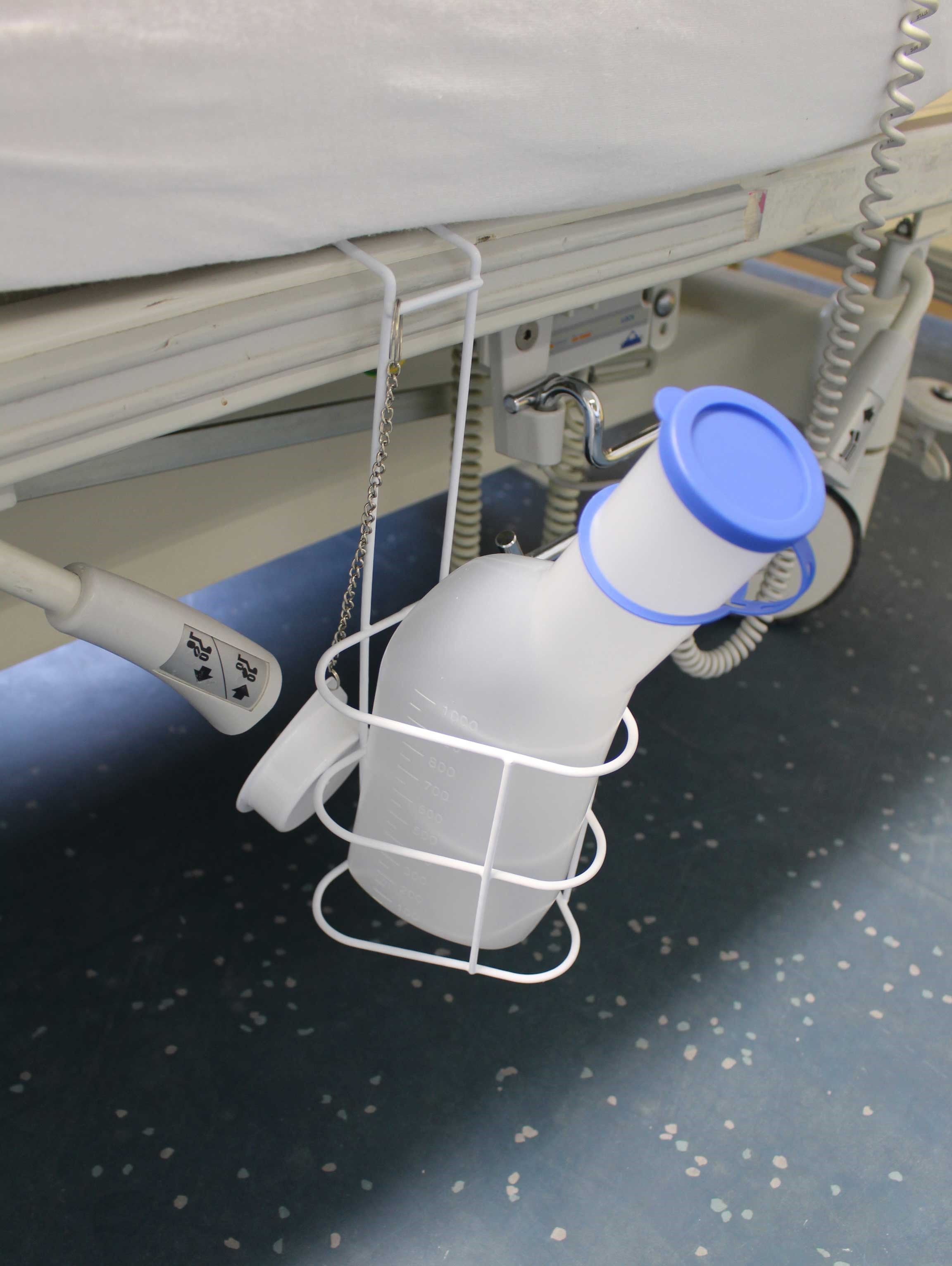 Urinflasche mit blauem Deckel in Halterung an einem Krankenhausbett. Milchiges Material. Halterung weiß.