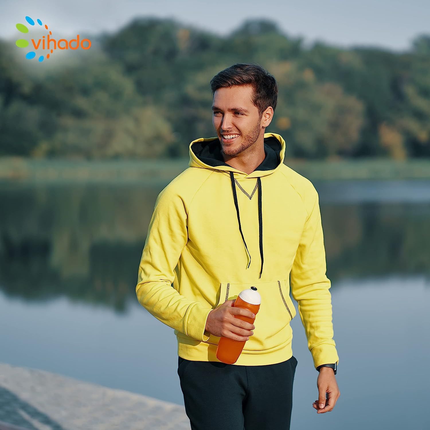 Ein Mann in gelbem Hoodie mit Wasserflasche. Hintergrund: See und Bäume. Der Mann lächelt.