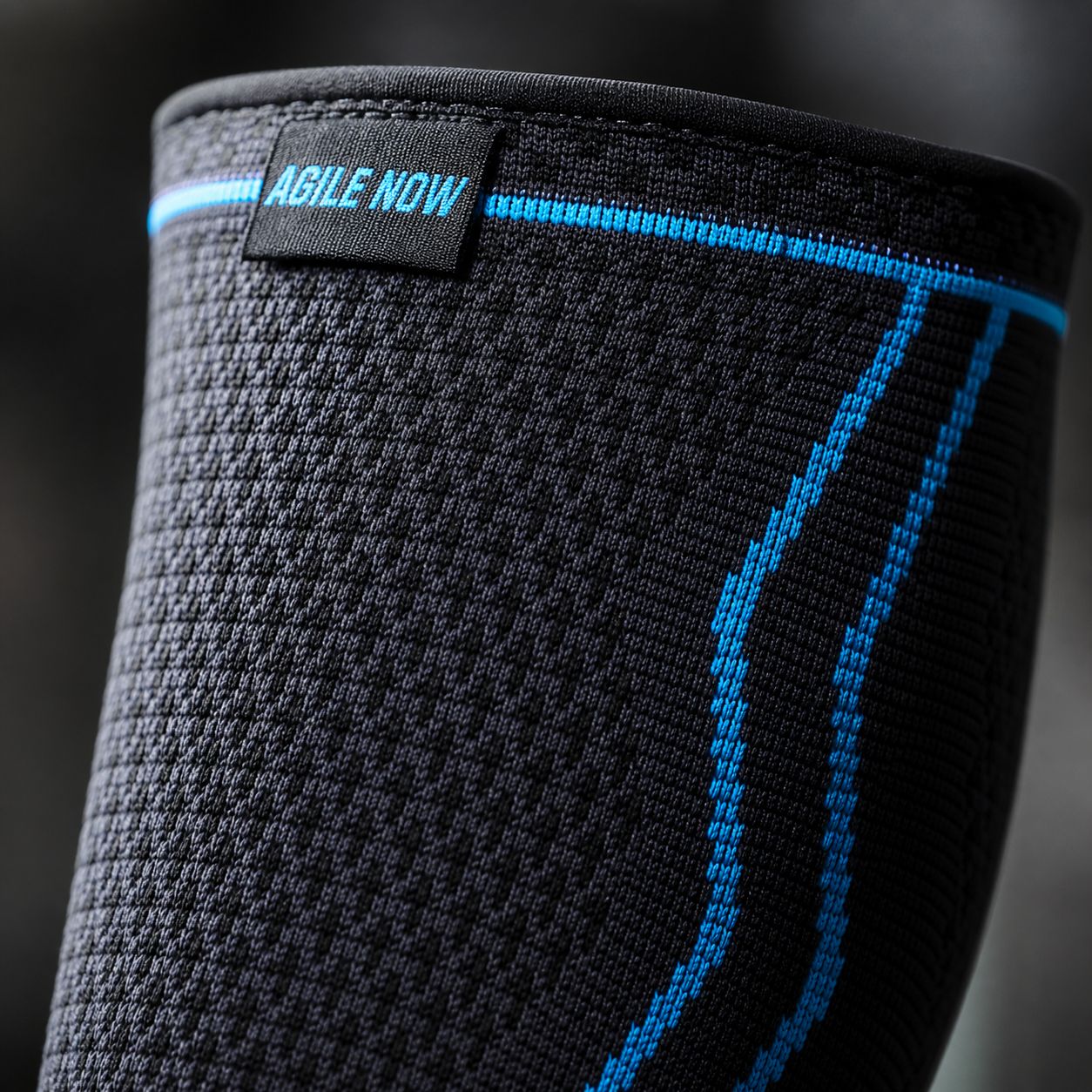 Nahaufnahme einer schwarzen Wadenbandage mit blauen Linien und dem Text AGILE NOW. Schwarzer Hintergrund.