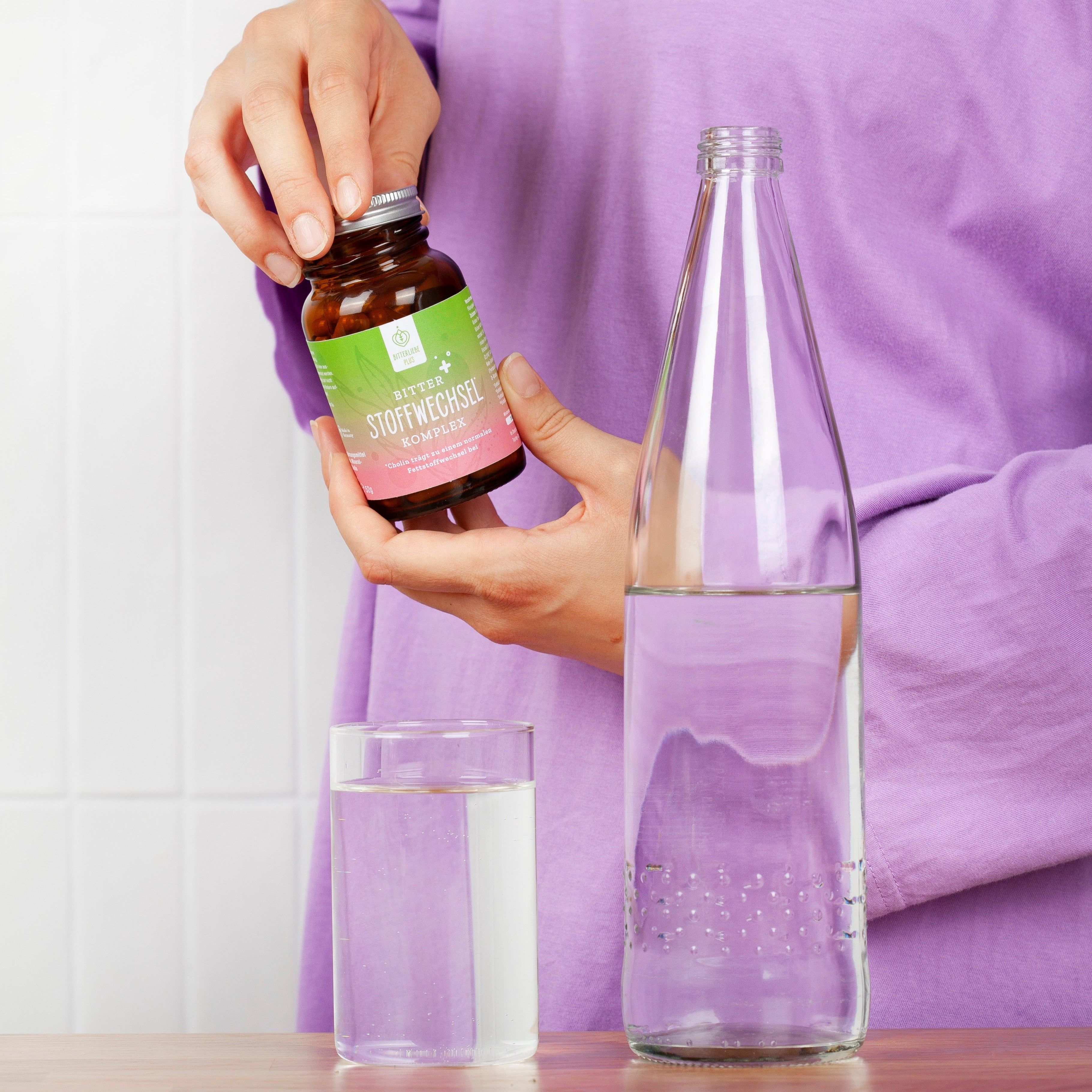 Person hält Flasche und Glas. Aufschrift: Bitter Stoffwechsel Komplex. Neben einer Glasflasche mit Wasser.