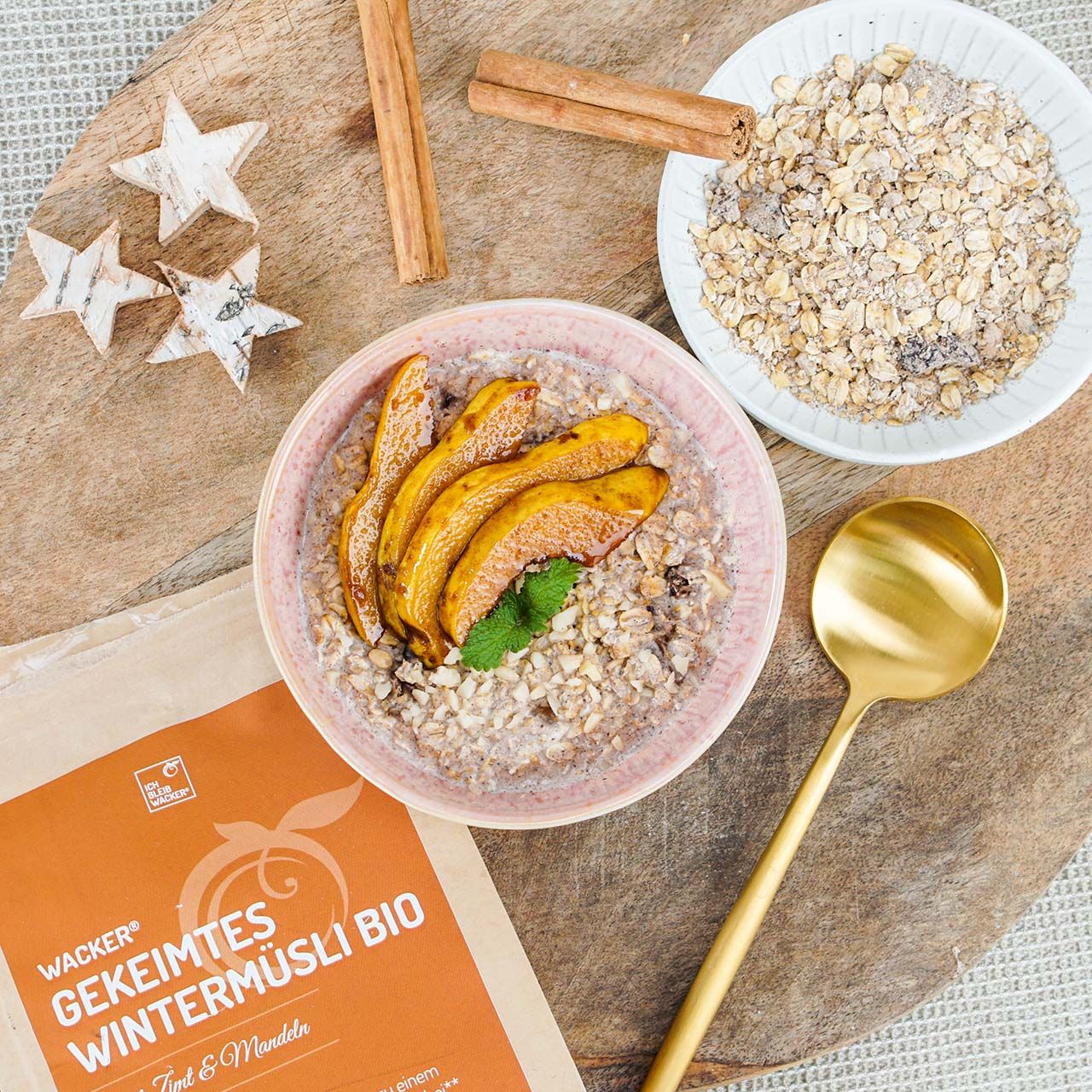 Müsli in Schale mit Apfelscheiben, Zimtstangen, Sternen und goldener Löffel. Packung Wacker Gekeimtes Wintermüsli Bio.