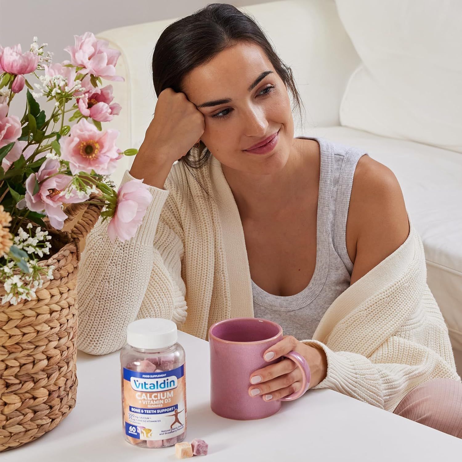 Frau sitzt mit einer Flasche VITALDIN Calcium + Vitamin D3 Gummies und einer Tasse. Aufschrift: Knochen- und Zahnunterstützung. Enthält 60 Gummies. Auf dem Tisch.