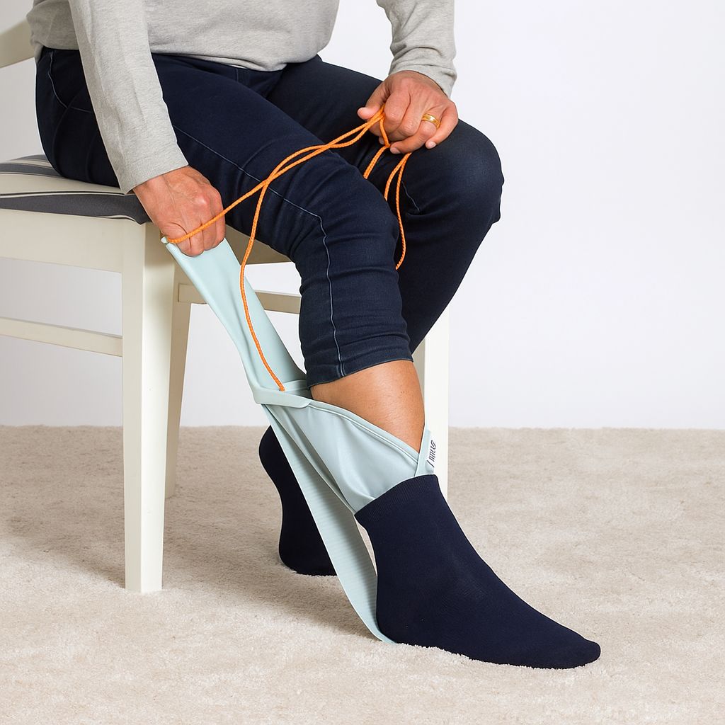 Person sitzt und benutzt ein hellblaues Hilfsmittel, um eine Socke anzuziehen. Orangefarbene Schnüre werden gehalten.