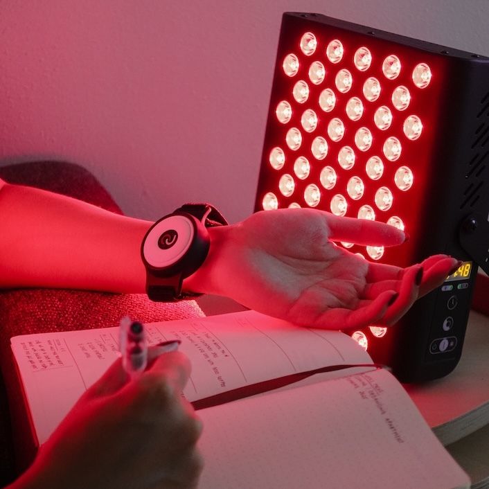 Person mit Armband und Lichttherapiegerät. Person schreibt in Notizbuch. Gerät strahlt rotes Licht.