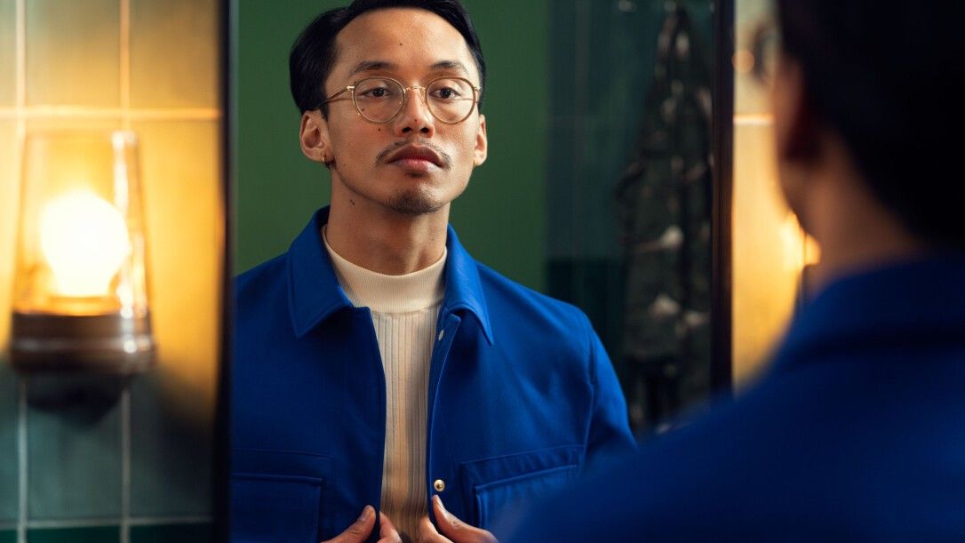 Ein Mann betrachtet sich im Spiegel. Er trägt eine Brille und eine blaue Jacke.