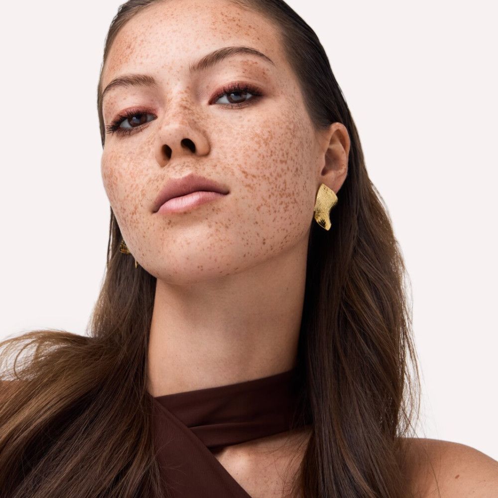 Porträt einer Frau mit Sommersprossen. Sie trägt goldene Ohrringe und rotes Augen-Make-up. Braunes Oberteil.