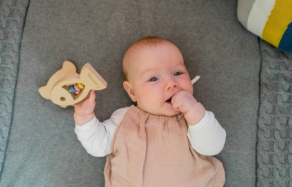 Baby liegt auf grauer Decke und hält einen Holz-Beißring in Fischform. Baby steckt Finger in den Mund.