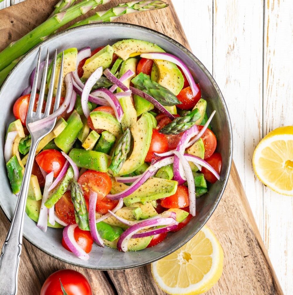 Salatschüssel mit Avocado, Tomaten, Zwiebeln und Spargel. Zitrone und Gabel daneben. Auf Holzbrett.