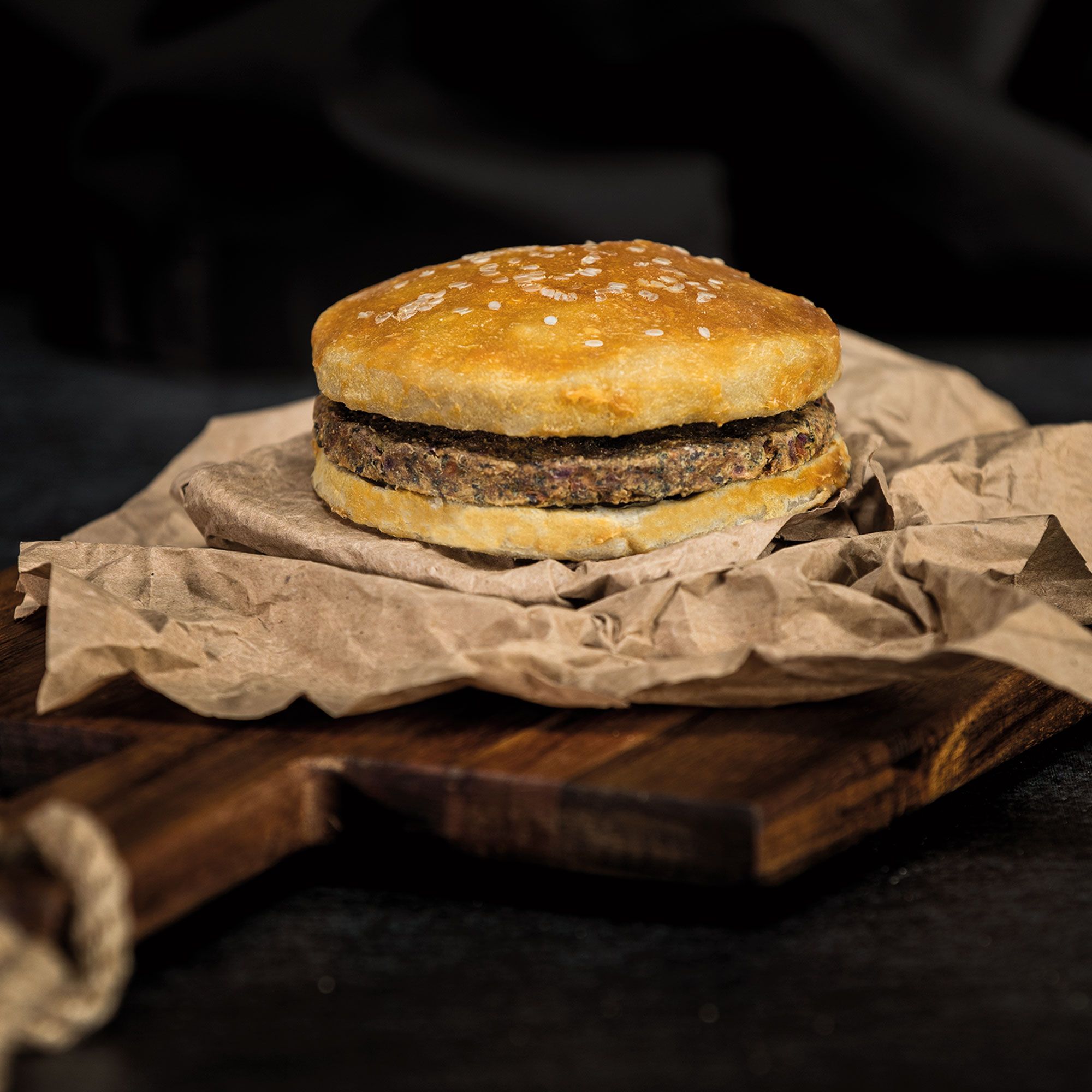 Hunde-Burger auf Holzbrett und Papier. Dunkles Brötchen mit Sesam, dunkles Fleisch.