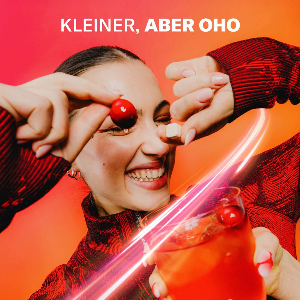 Person hält Kirsche und Würfel. Ein Glas mit roter Flüssigkeit. Text: Kleiner, aber Oho. Wasserdrop® Logo.