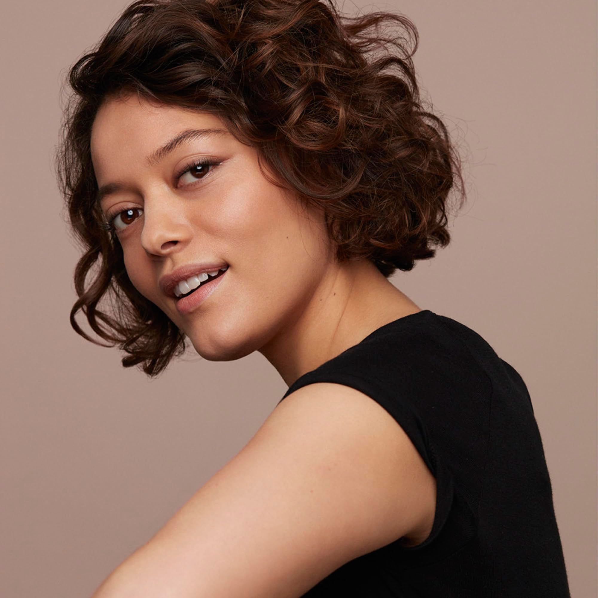 Porträt einer Frau mit lockigem, braunem Haar. Sie trägt ein schwarzes Oberteil und lächelt in die Kamera.