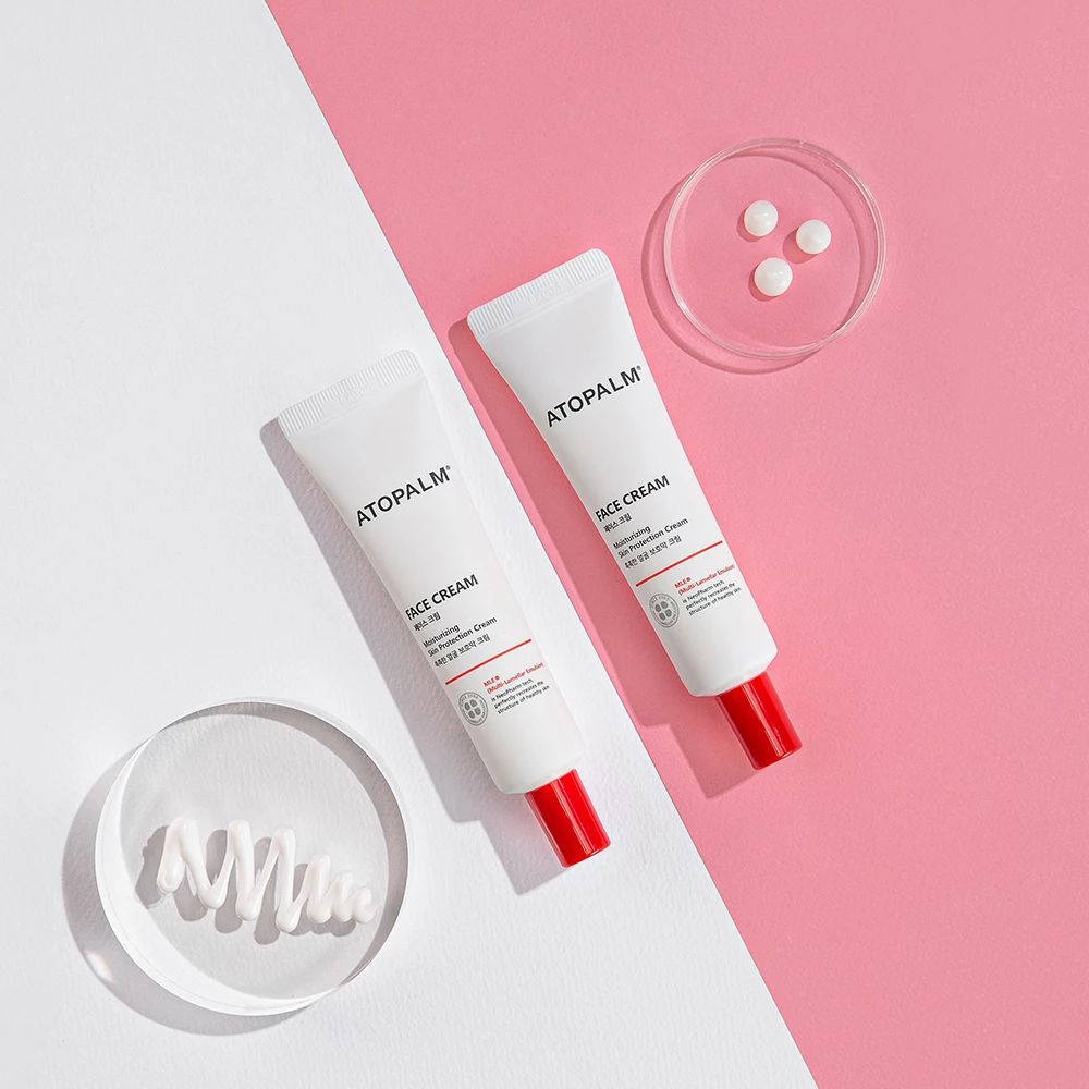 Zwei weiße Tuben mit rotem Verschluss. Aufschrift: ATOPALM FACE CREAM. Zusätzliche Elemente: transparente Platte, Kügelchen.