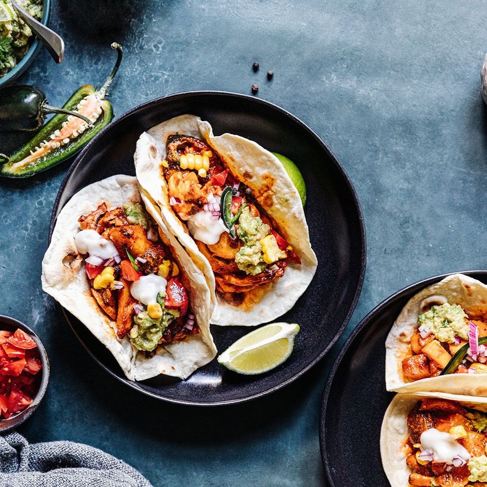 Tacos gefüllt mit Gemüse und Sauce. Limettenspalten. Auf schwarzem Teller. Weitere Schalen mit Zutaten.