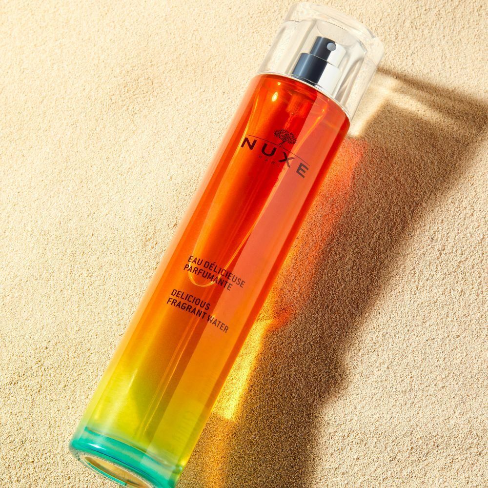 Eine Flasche mit farbigem Inhalt auf Sand. Auf der Flasche steht NUXE Sun und der Text Eau Délicieuse Parfumante.
