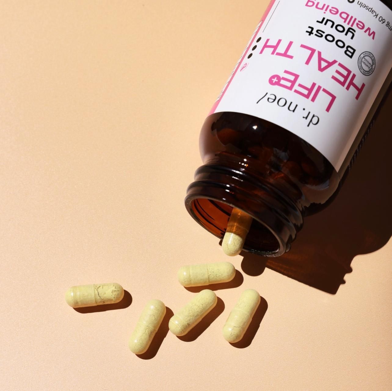 Braune Glasflasche mit schwarzem Deckel und Etikett. Kapseln liegen daneben. Aufschrift: Dr. Noel Life+ Health, Boost your wellbeing. Nahrungsergänzungsmittel.