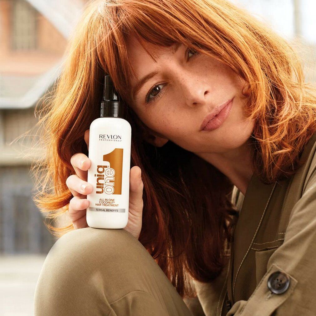 Frau hält ein weißes Spray mit schwarzem Sprühkopf. Aufschrift: Revlon Professional Uniq One All in One Coconut Hair Treatment.
