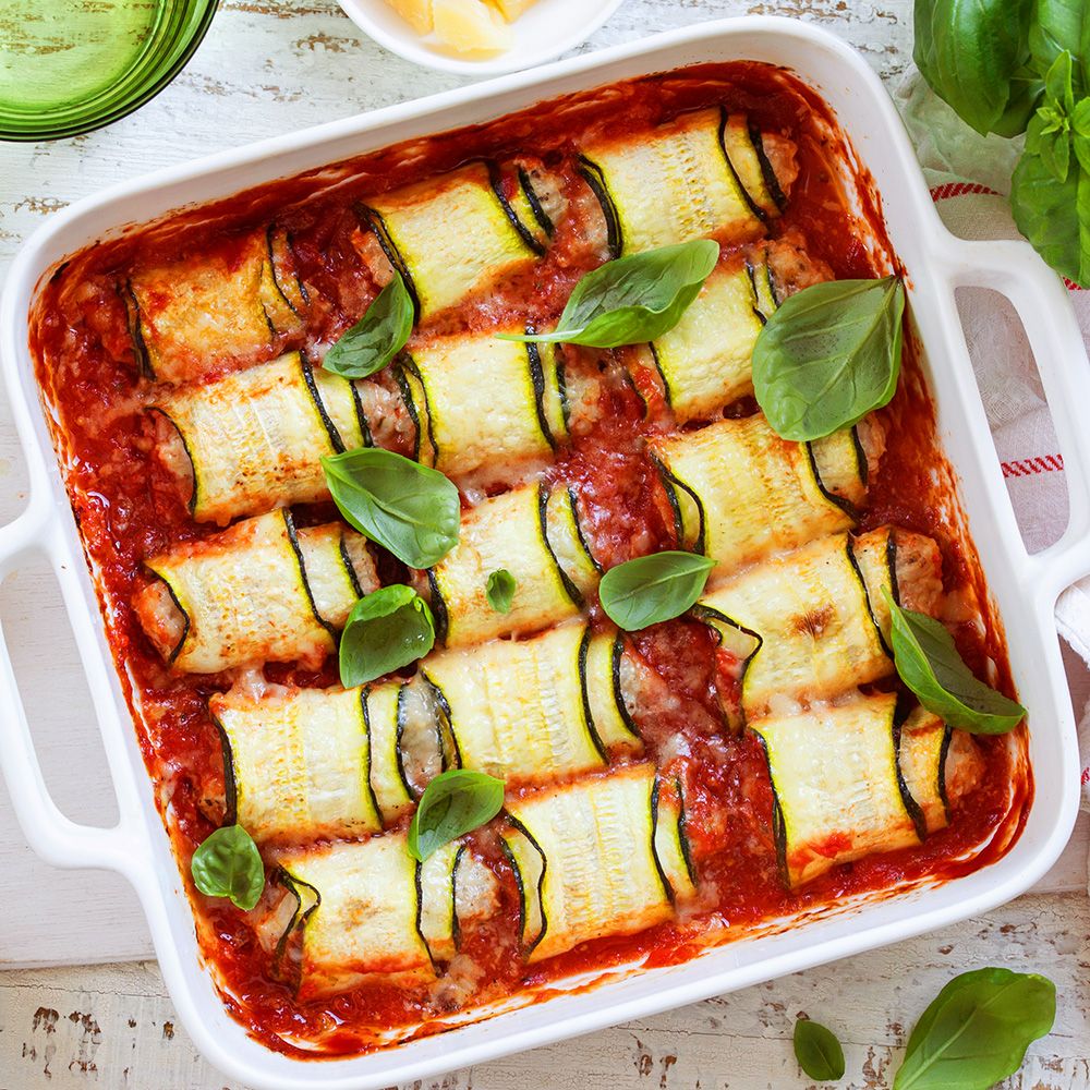 Auflaufform mit Zucchiniröllchen in Tomatensauce. Basilikumblätter als Dekoration. Helle Holzoberfläche.