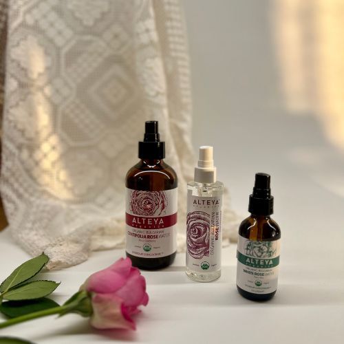 Drei Sprühflaschen mit Rosenwasser. Aufschrift: Alteya Organics. USDA Organic-Zertifizierung. Eine Rose liegt davor.