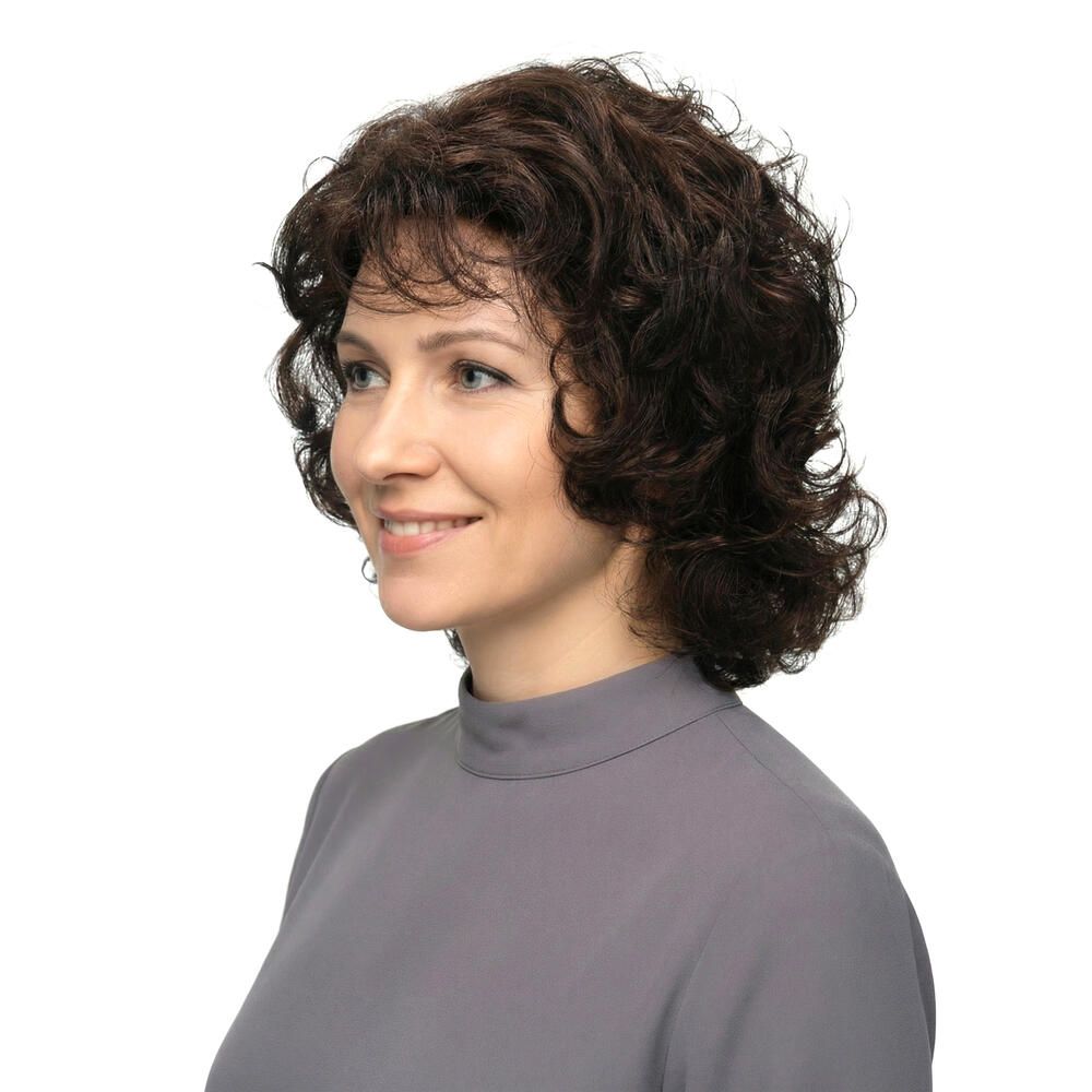 Frau mit brauner, lockiger Perücke. Seitenansicht, lächelnd. Graues Oberteil.