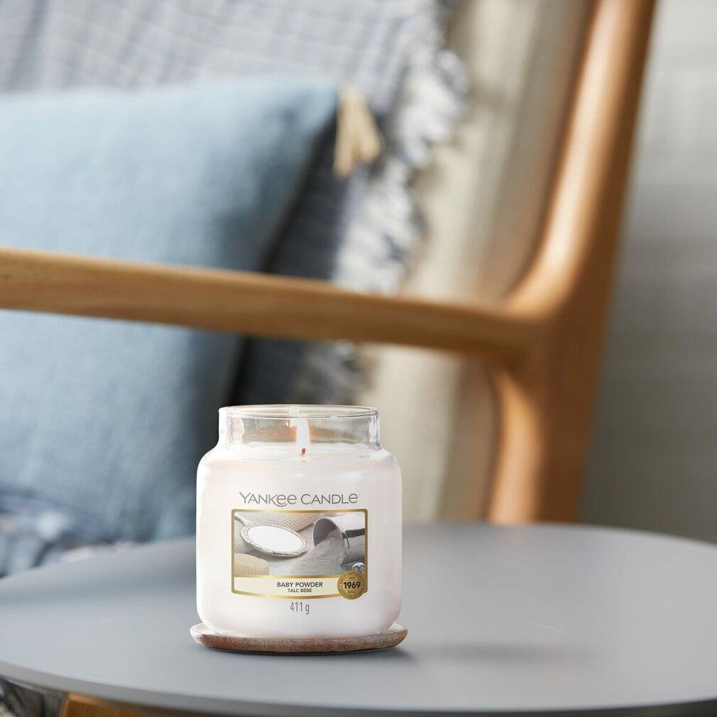 Yankee Candle Baby Powder Duftkerze auf einem Tisch. Im Hintergrund Stuhl und Kissen. Gewicht: 411g.