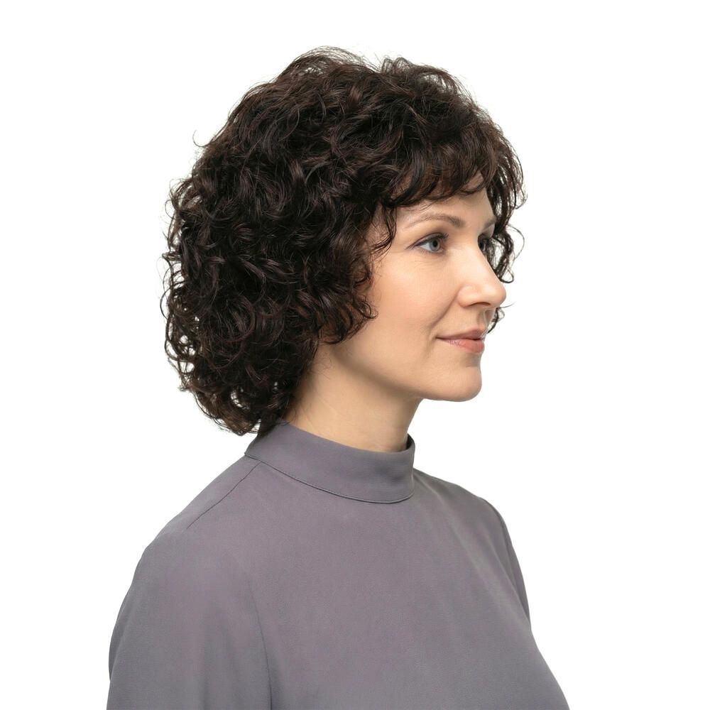 Frau mit brauner, lockiger Perücke. Seitenansicht, lächelnd. Graues Oberteil.