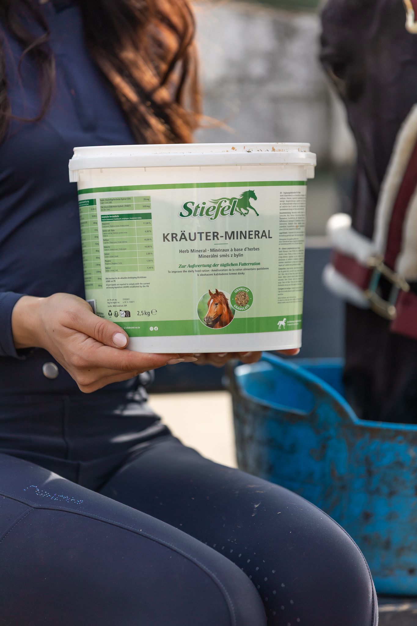Weißer Eimer mit grünem Etikett. Aufschrift: Stiefel Kräuter-Mineral. Ein Pferd wird gefüttert. 2,5 kg.