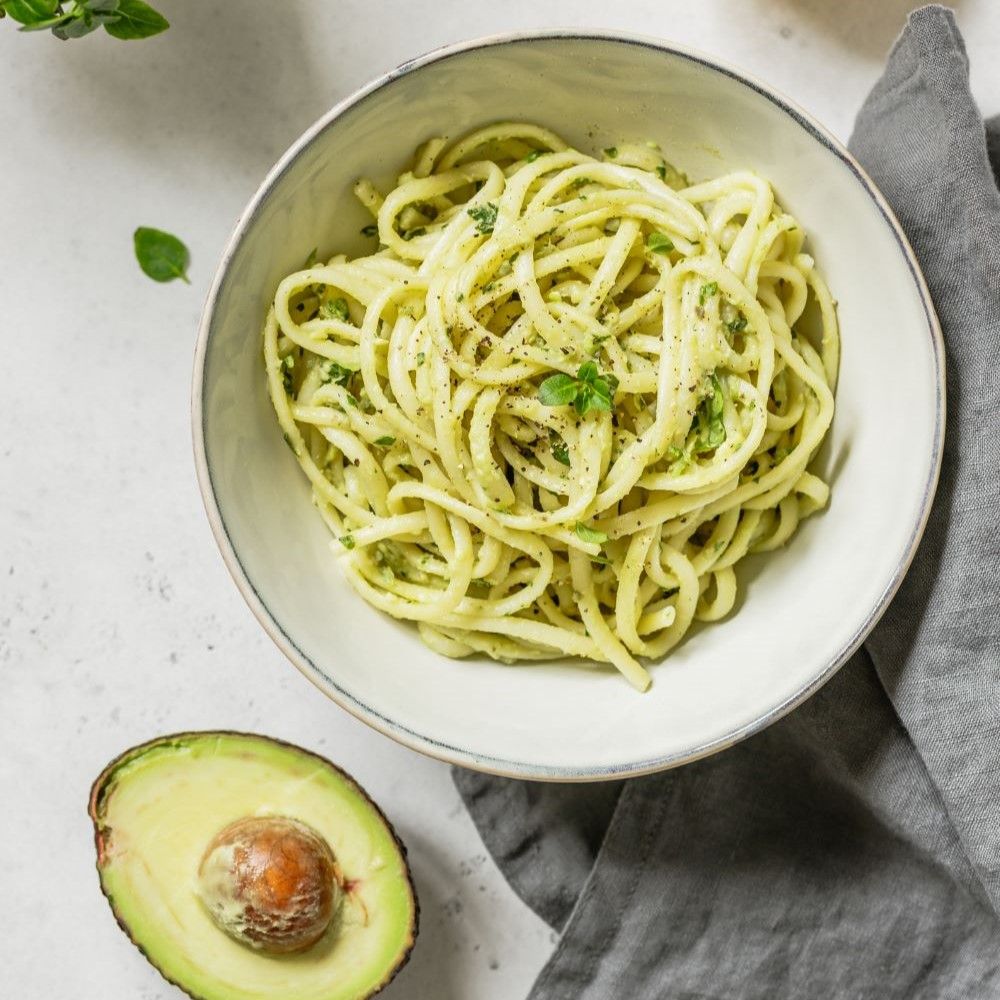 Teller mit Pasta, Pesto und Basilikum. Daneben eine halbe Avocado. Auf hellem Untergrund.