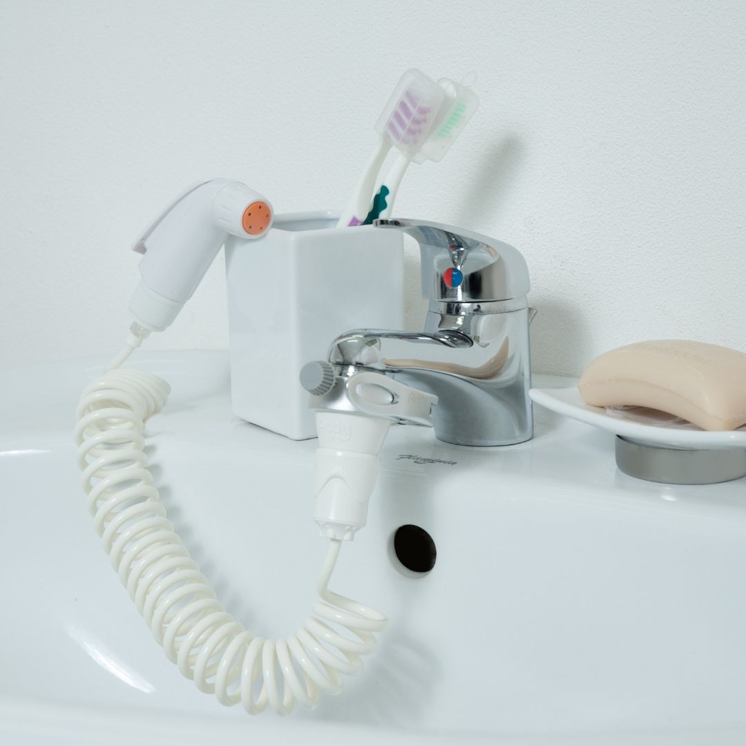 Doccetta bidet portatile vicino al lavandino. Doccetta e adattatore collegati al rubinetto. Lavandino bianco.