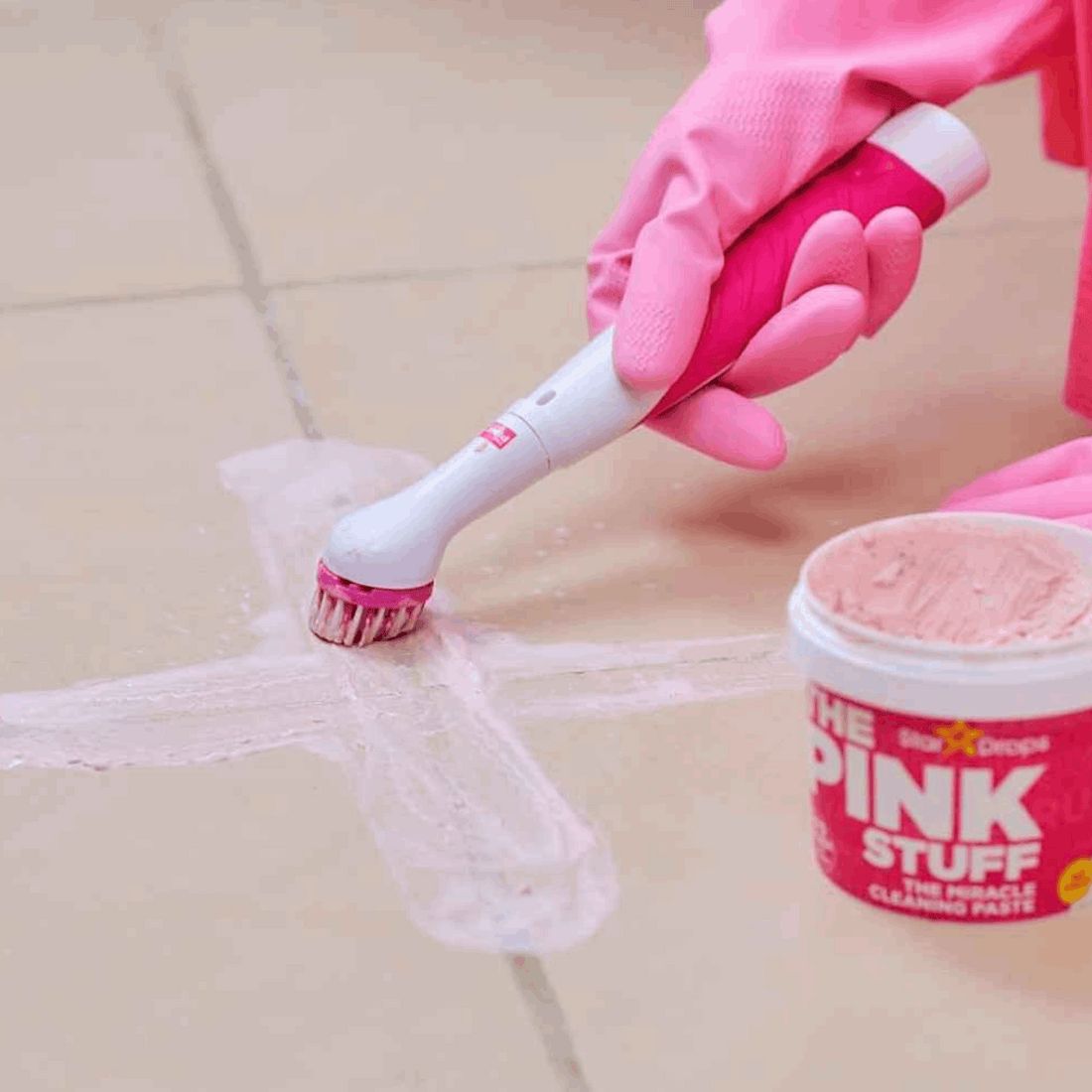 Person hält THE PINK STUFF Sonic Scrubber. Rosa Handschuhe. Daneben ein Tiegel THE PINK STUFF Reinigungs-Paste. Reinigung auf Fliesen.