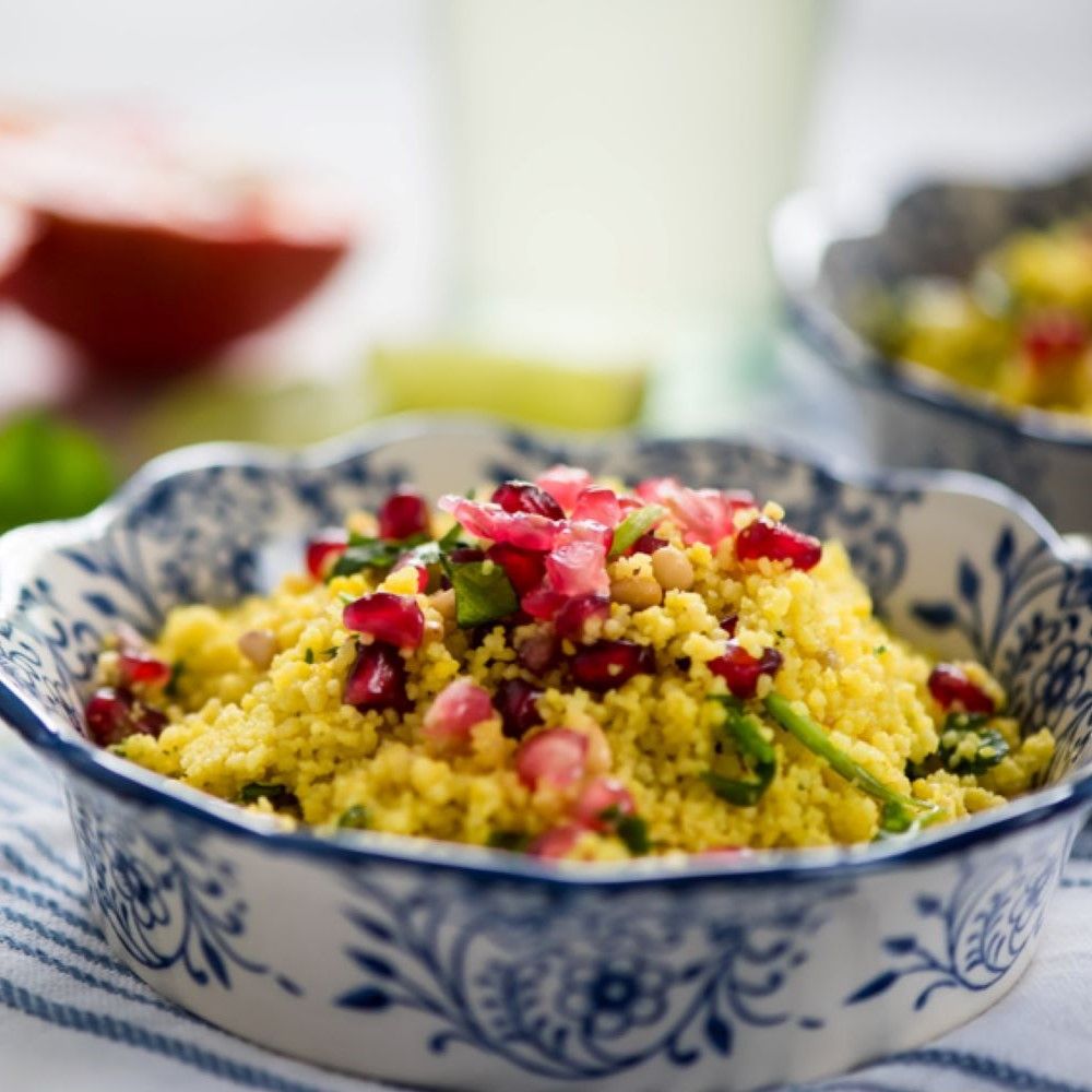 Zwei Schalen mit Couscous, Granatapfelkernen und Kräutern. Blaue und weiße Musterung. Auf gestreiftem Tuch.