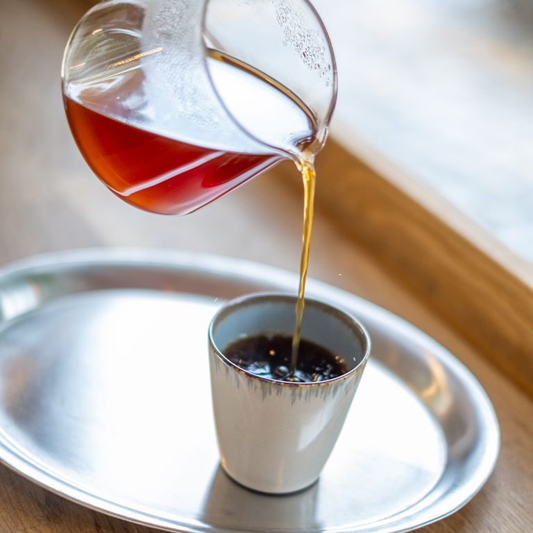 Kaffee wird in eine Tasse gegossen. Silberne Untertasse. Helle Holztischplatte.