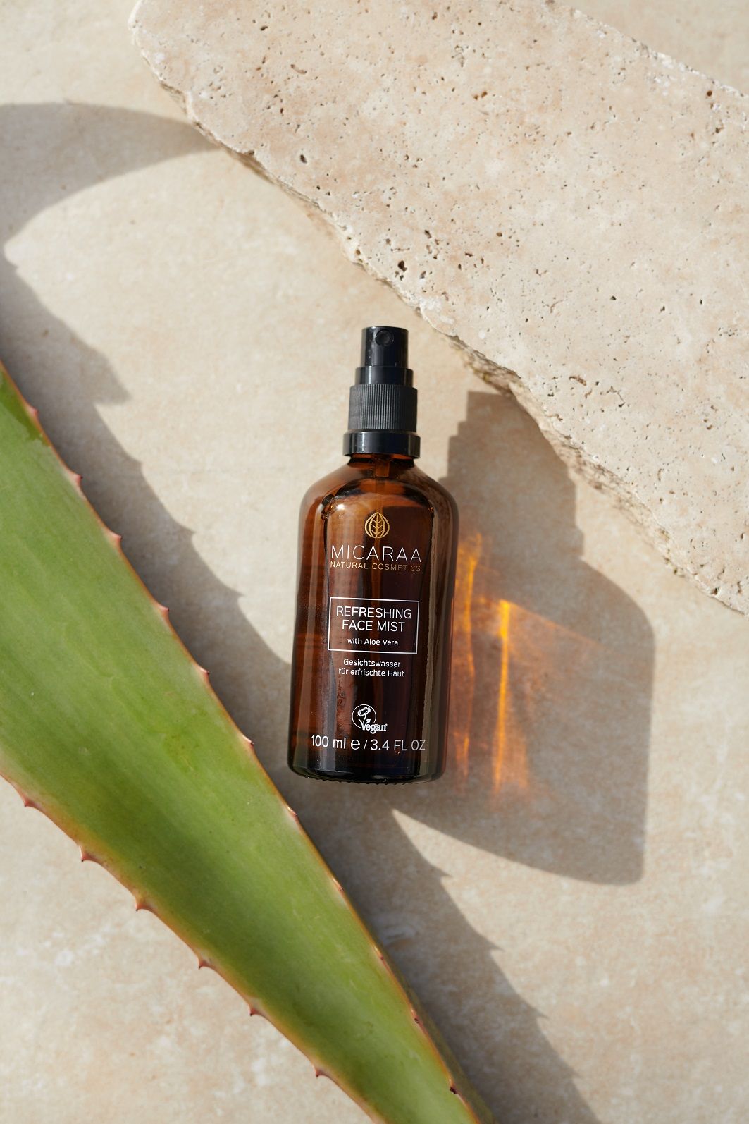 Braune Glasflasche mit Sprühkopf. Beschriftung: MICARAA, Refreshing Face Mist, mit Aloe Vera. Vegan-Siegel. Auf Stein und Aloe-Blatt.