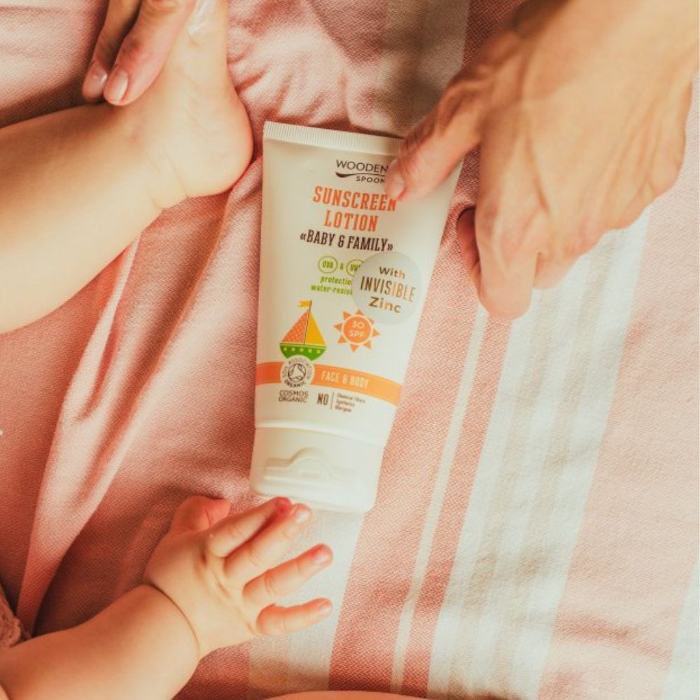 Sonnencreme-Tube auf rosa Stoff. Hände berühren die Tube und ein Babyfuß. Aufschrift: SUNSCREEN LOTION «BABY & FAMILY».