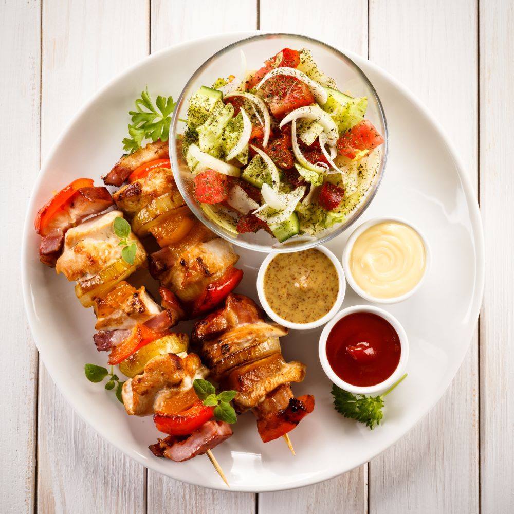 Teller mit Hähnchen-Spießen, Salat und Saucen. Gegrilltes Fleisch und Gemüse auf Spießen. Salat in Glasschale, Saucen in kleinen Schalen.